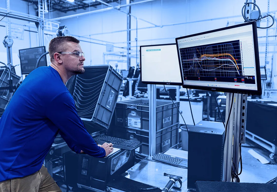 Worker wearing safety glasses operating a computer at a factory testing station while viewing line graphs on two monitors.