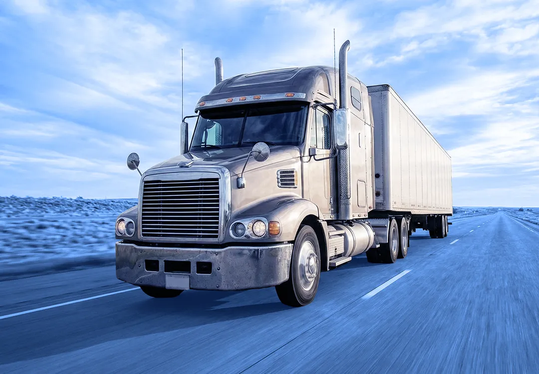 Semi truck pulling a box trailer driving along a paved highway through an open landscape under a cloudy sky.