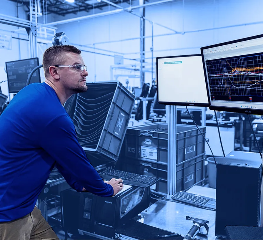 Worker wearing safety glasses operating a computer at a testing station while viewing data charts on two monitors inside a factory floor workspace.