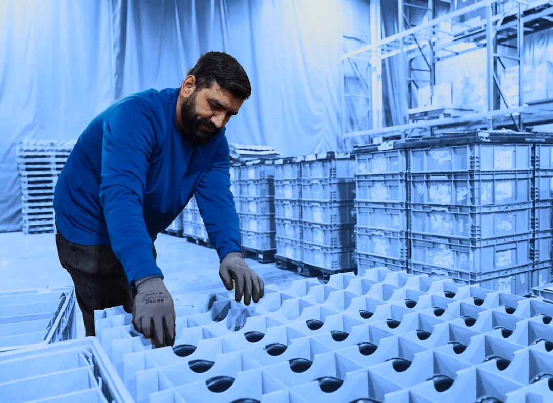 Worker sorting plastic containers inside a warehouse facility with stacked returnable packaging in the background.