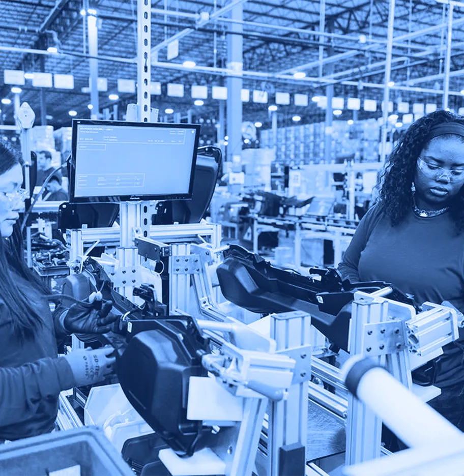 Two workers wearing safety glasses assembling plastic components at a workstation with fixtures, tools, and a monitor inside a warehouse.