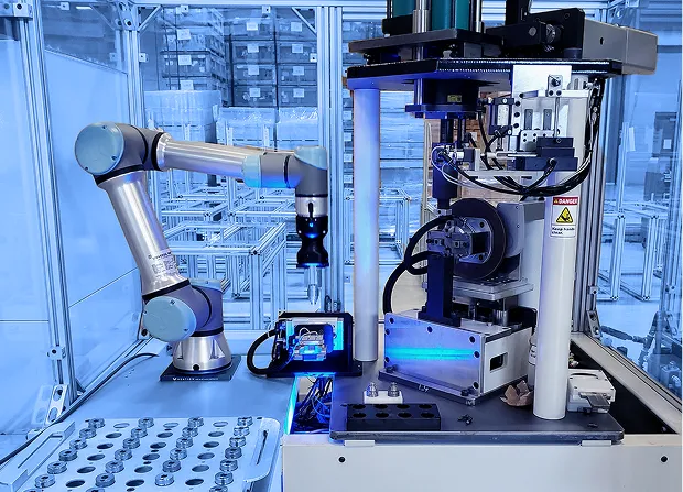 Robotic arm positioned beside a vertical press assembly inside an enclosed automation cell with fixtures, sensors, and metal parts arranged on a work surface.