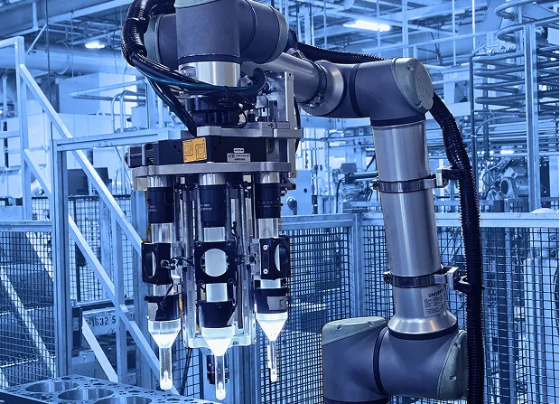 Multi-tool robotic arm positioned above a metal engine block inside a fenced manufacturing cell with lighting, cables, and structural supports visible. 
