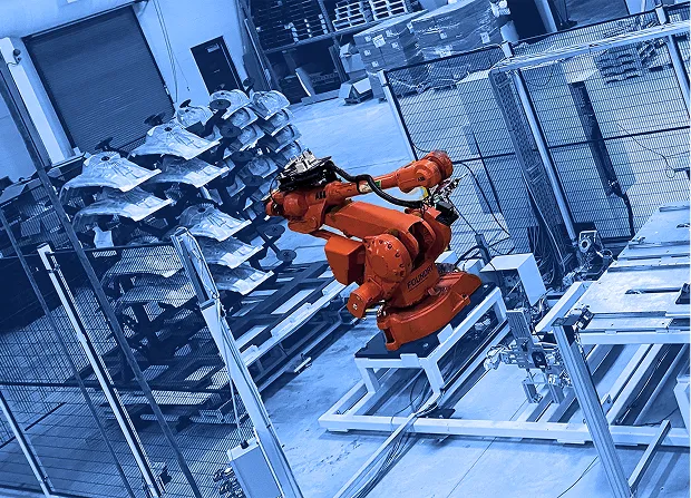 Two industrial robotic arms positioned inside a fenced automation cell with fixtures, worktables, and stacked parts visible in a factory space.