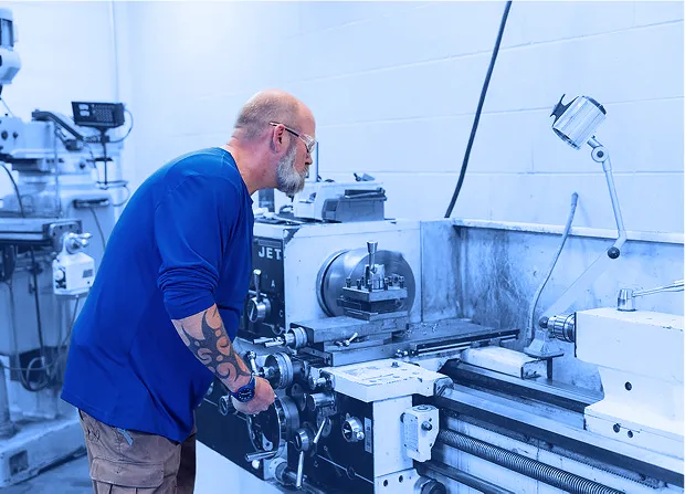 Worker wearing safety glasses operating a metal lathe inside a machine shop with tooling and controls visible.