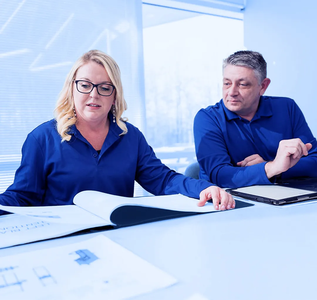 Two office workers seated at a table reviewing printed documents and a laptop during a meeting in a conference room.