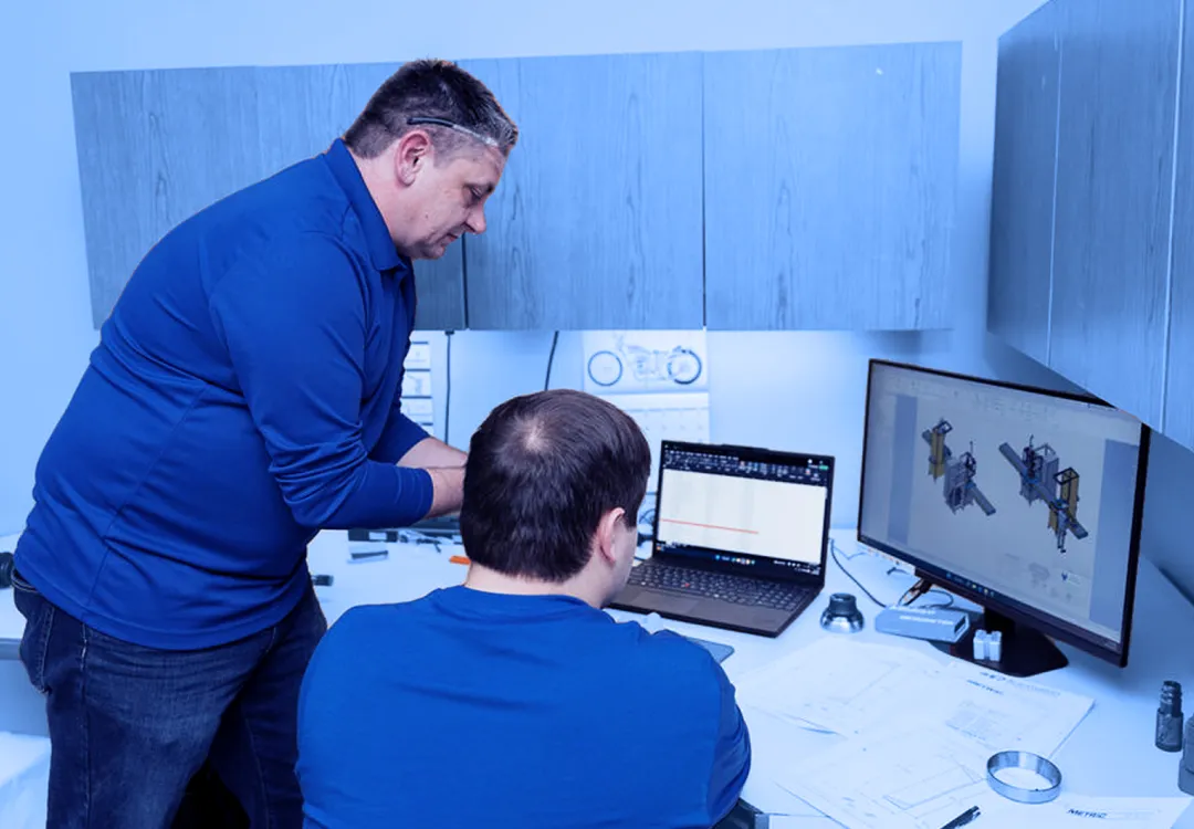 Two office workers reviewing technical drawings on a computer monitor and laptop at a desk with printed plans and tools visible.  