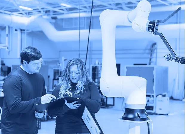 Two workers wearing safety glasses reviewing documents while standing beside a collaborative robotic arm inside a factory workspace. 