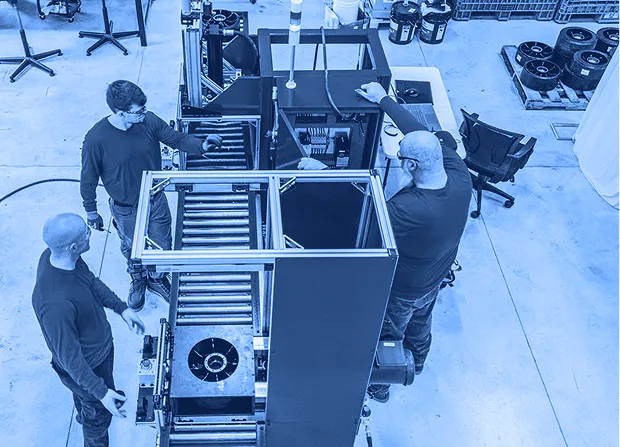 Three workers standing around a blue industrial machine with a roller conveyor inside a factory workspace, viewed from above. 