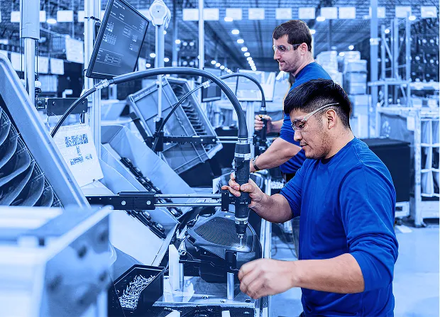 Worker uses a mounted power screwdriver to fasten components on a black plastic assembly at a workstation while another worker stands behind monitoring the process.