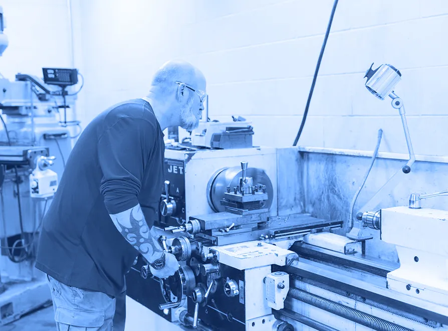 Worker wearing safety glasses operating a metal lathe inside a machine shop with tooling and controls visible.