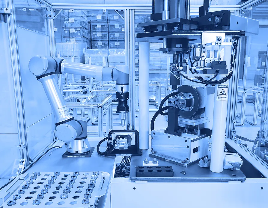 Robotic arm positioned beside a vertical press assembly inside an enclosed automation cell with fixtures, sensors, and metal parts arranged on a work surface.  