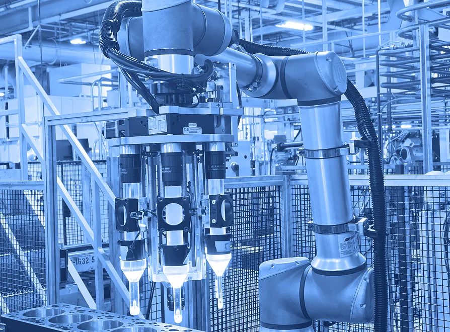 Multi-tool robotic arm positioned above a metal engine block inside a fenced manufacturing cell with lighting, cables, and structural supports visible.