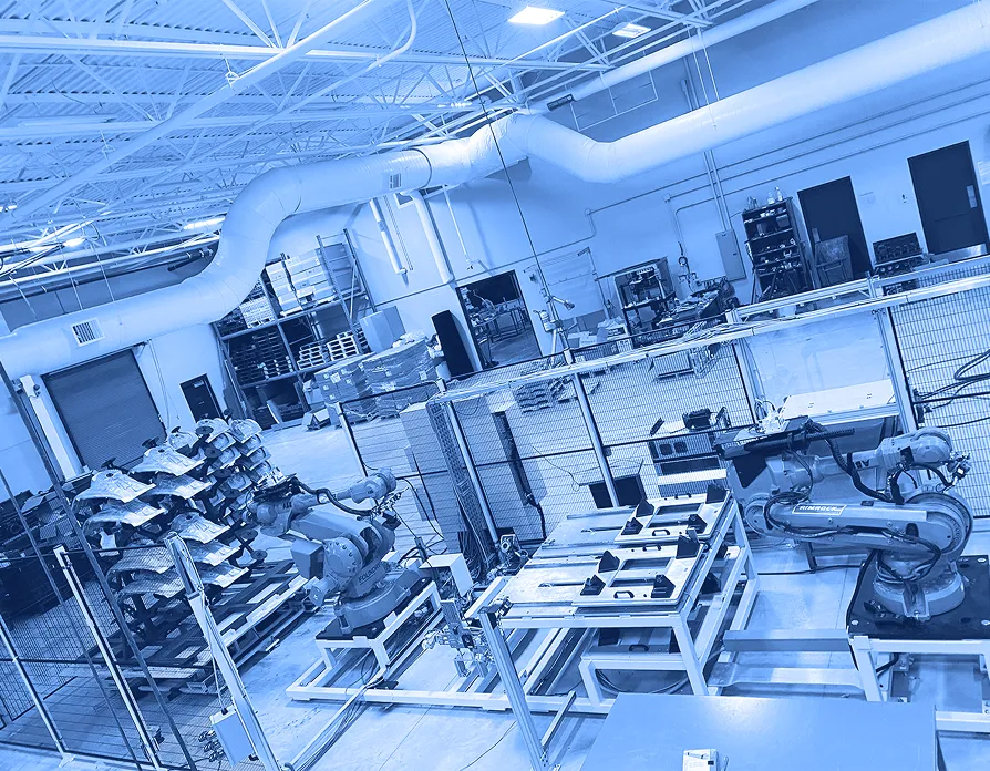 Two industrial robotic arms positioned inside a fenced automation cell with fixtures, worktables, and stacked parts visible in a factory space.