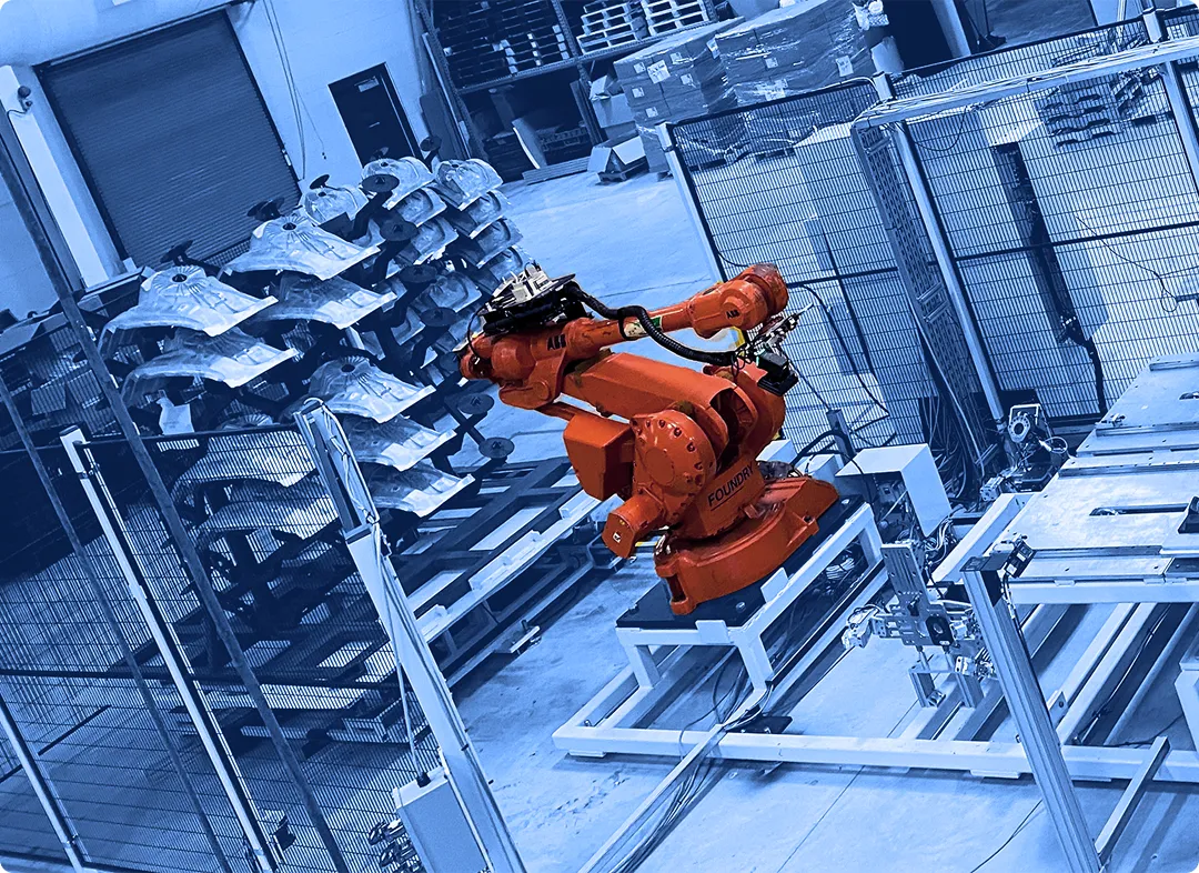 Two industrial robotic arms positioned inside a fenced automation cell with fixtures, worktables, and stacked parts visible in a factory space.