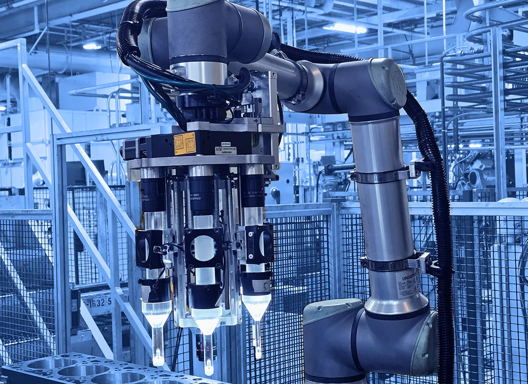 Multi-tool robotic arm positioned above a metal engine block inside a fenced manufacturing cell with lighting, cables, and structural supports visible.