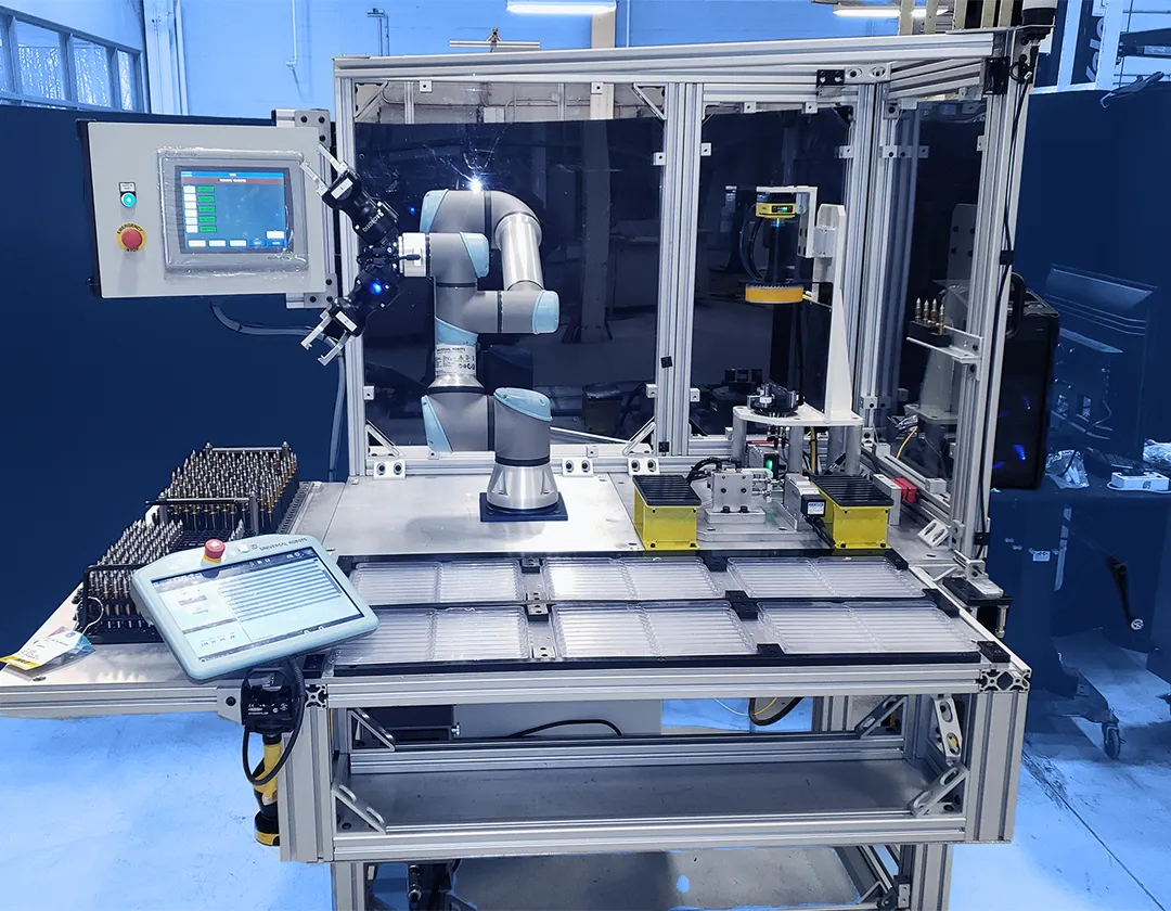 Collaborative robotic arm mounted inside an enclosed automation cell with a touchscreen control panel, fixtures, and work surfaces arranged around the station