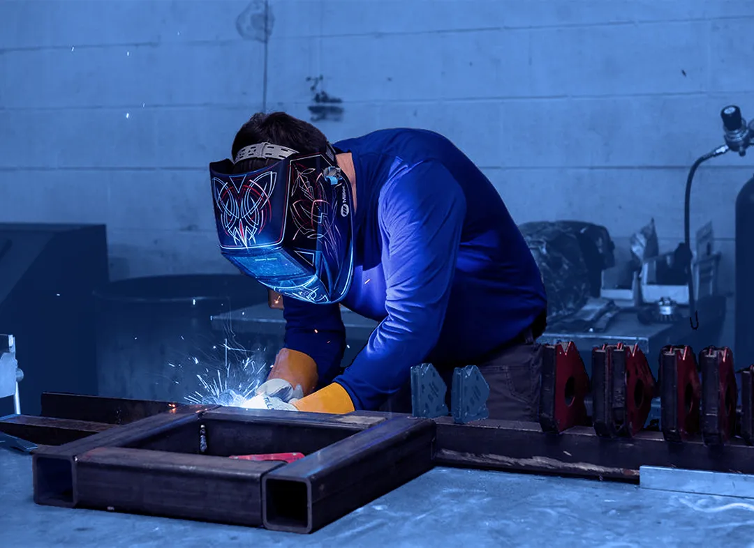 Worker wearing a welding helmet and gloves welding a metal frame on a workbench with tools and metal components nearby.
