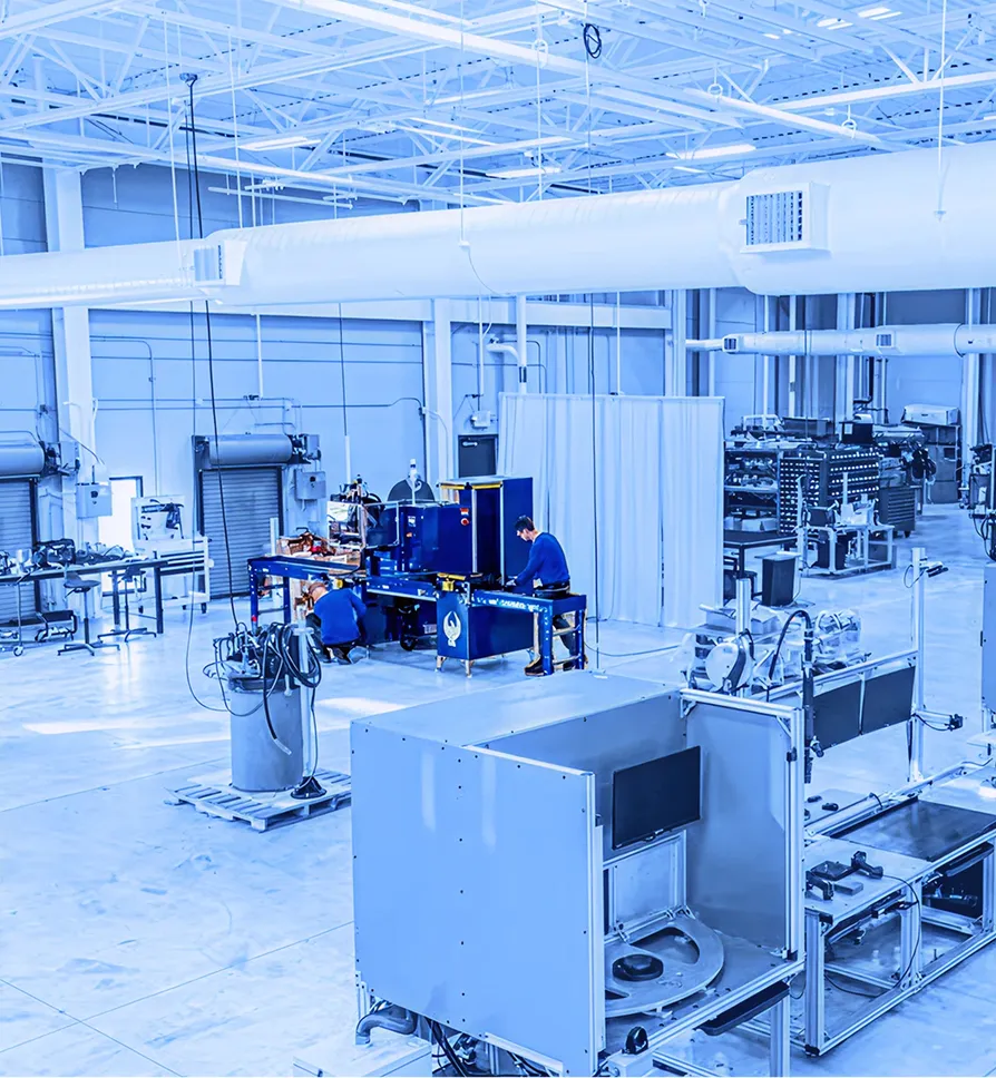 Wide view of a factory floor with multiple industrial machines, workstations, and workers spaced across an open manufacturing area under a high ceiling.