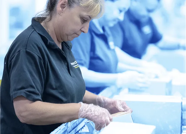 Three workers wearing gloves stand along a conveyor line, manually handling and closing small cardboard boxes in a warehouse or packaging facility.