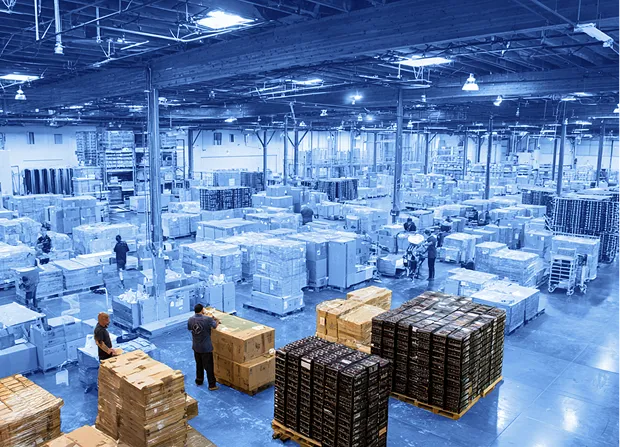 Side overhead view of a large warehouse interior with wooden ceiling beams, showing rows of pallets stacked with cardboard boxes and wrapped goods, workers moving between pallets, and open aisles across a concrete floor under overhead lighting.