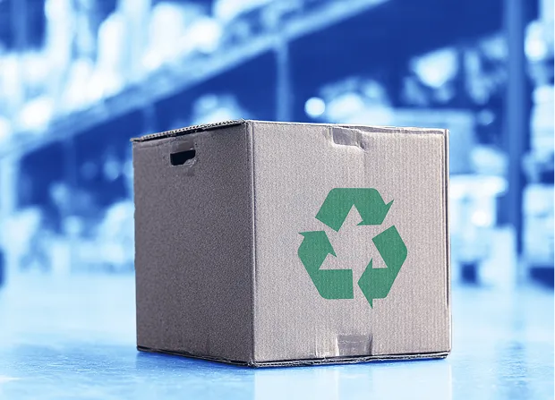 A cardboard box with a green recycling symbol printed on the side sits on a warehouse floor, with storage racks and stacked pallets visible in the background.