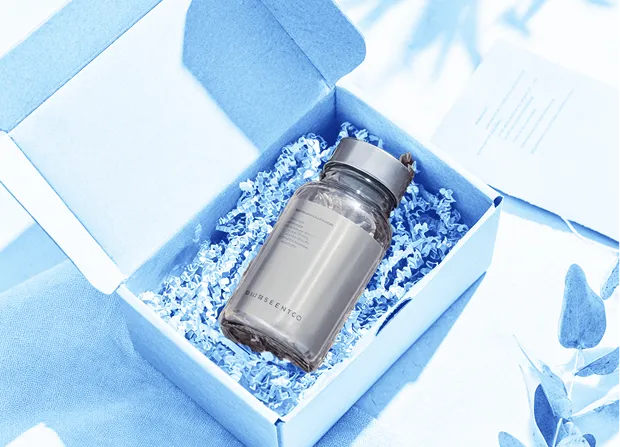 An open cardboard box contains a glass jar with a metal lid resting in shredded paper fill, with a folded paper card and plant leaves visible nearby.