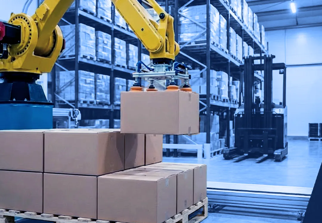 A yellow robotic arm uses suction cups to lift a cardboard box from a pallet stacked with boxes inside a warehouse, with storage racks and a forklift visible in the background.