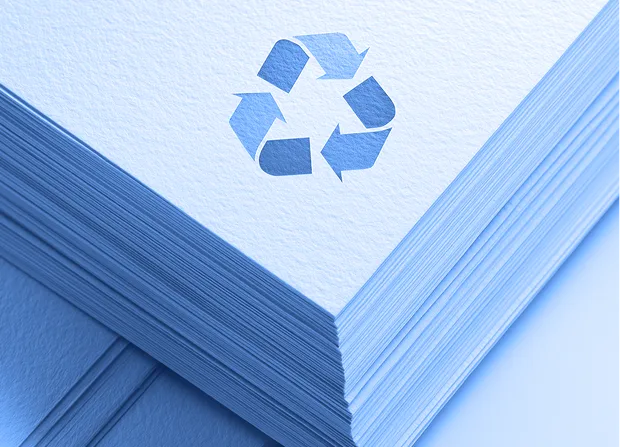 A stack of white paper sheets viewed from the corner, with a green recycling symbol printed on the top sheet.