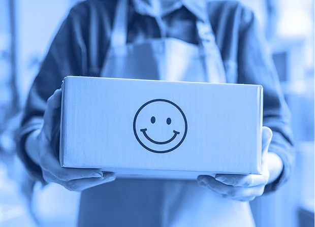 A person wearing an apron holds a rectangular cardboard box with a printed smiley face on the front.