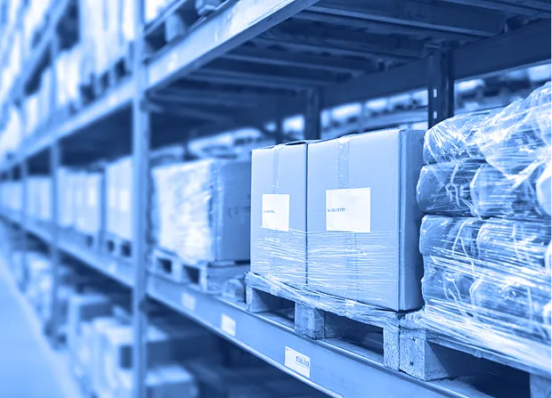 Cardboard boxes wrapped in clear plastic sit on wooden pallets on metal shelving inside a warehouse aisle.