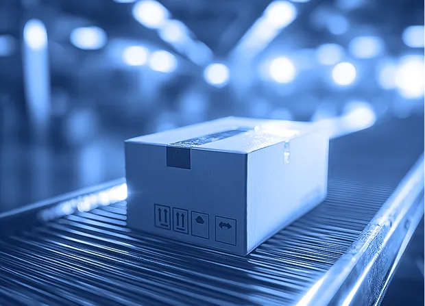 A sealed cardboard box moves along a metal conveyor belt inside a warehouse with overhead lights visible in the background.