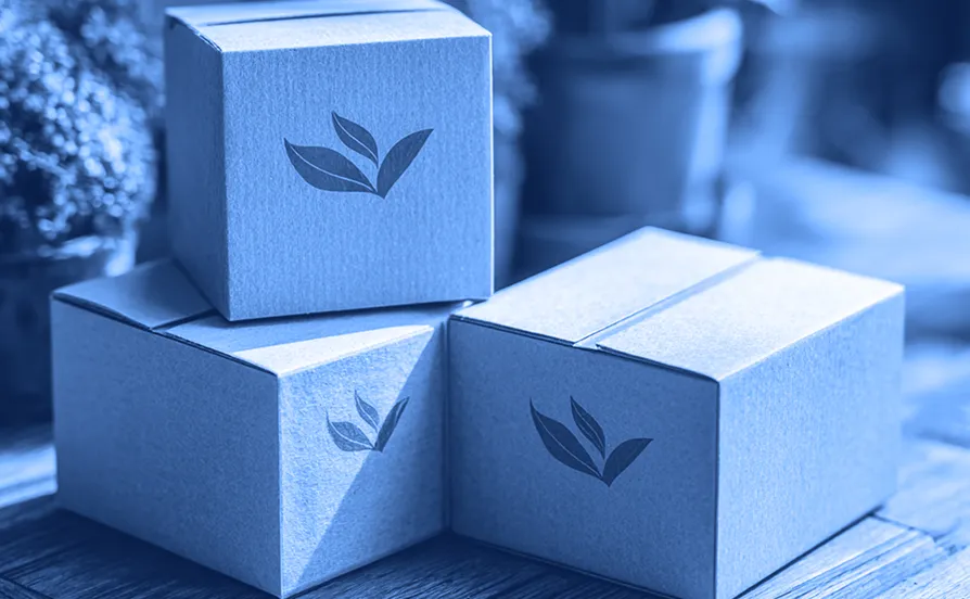 Three small brown cardboard boxes stacked on a wooden surface, each printed with a green leaf symbol, with potted plants blurred in the background.