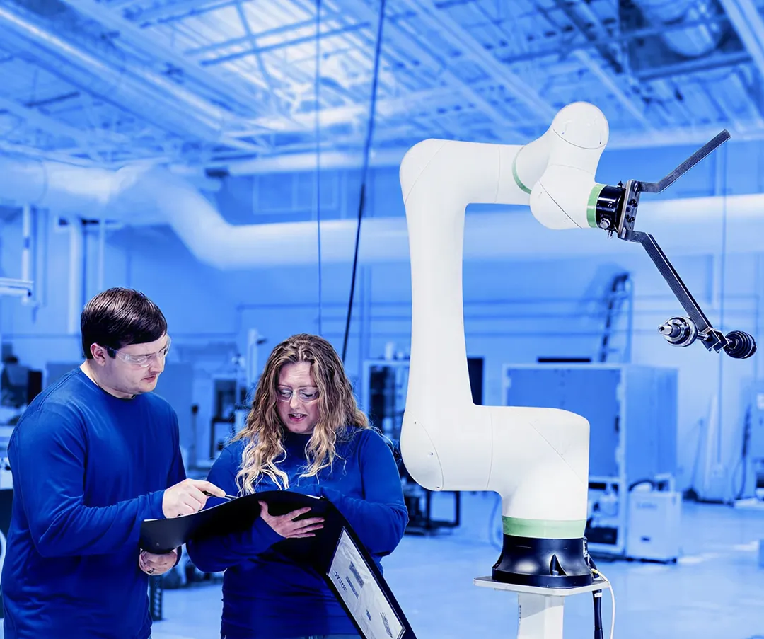 Two workers wearing safety glasses reviewing documents while standing beside a collaborative robotic arm inside a factory workspace.