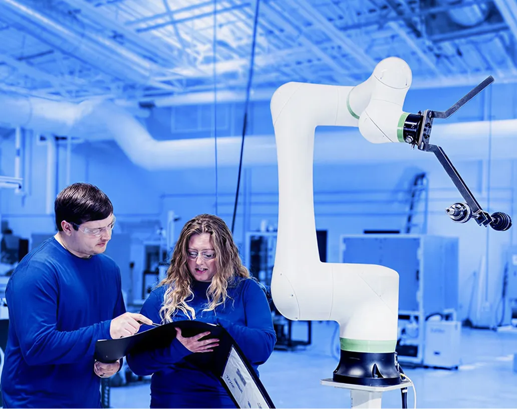 Two workers wearing safety glasses reviewing documents while standing beside a collaborative robotic arm inside a factory workspace.