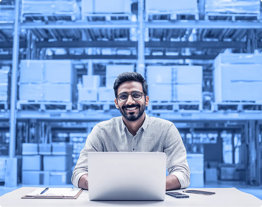 A person sits at a table using a laptop in a warehouse, with shelves of stacked cardboard boxes and pallets visible in the background.