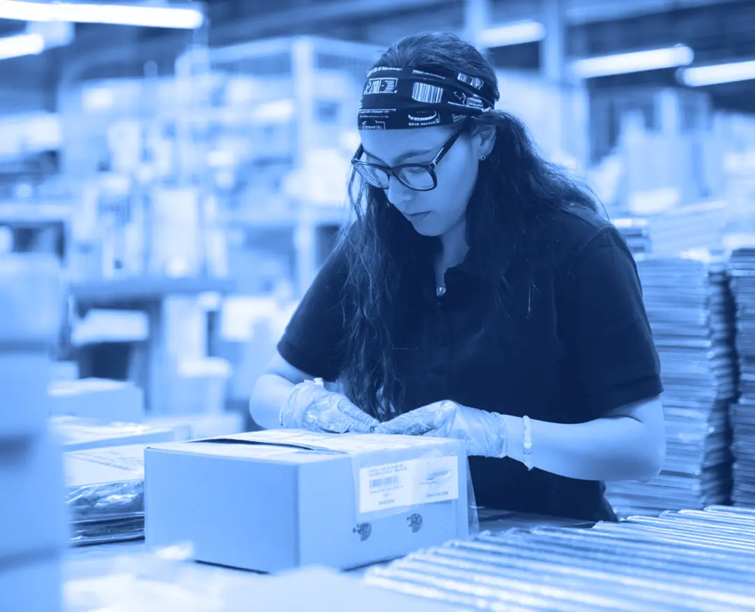 A worker wearing gloves applies a shipping label to a cardboard box on a packing table inside a warehouse, with stacked boxes and shelving visible in the background.