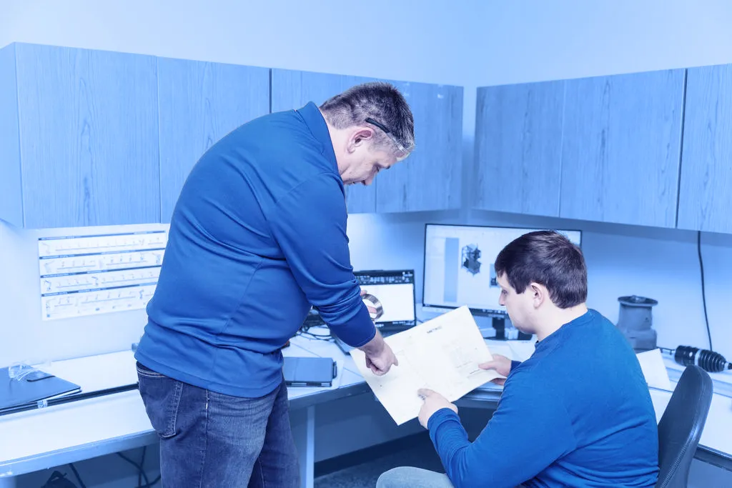 Two employees review printed engineering drawings at a desk with computer monitors displaying 3D component models.