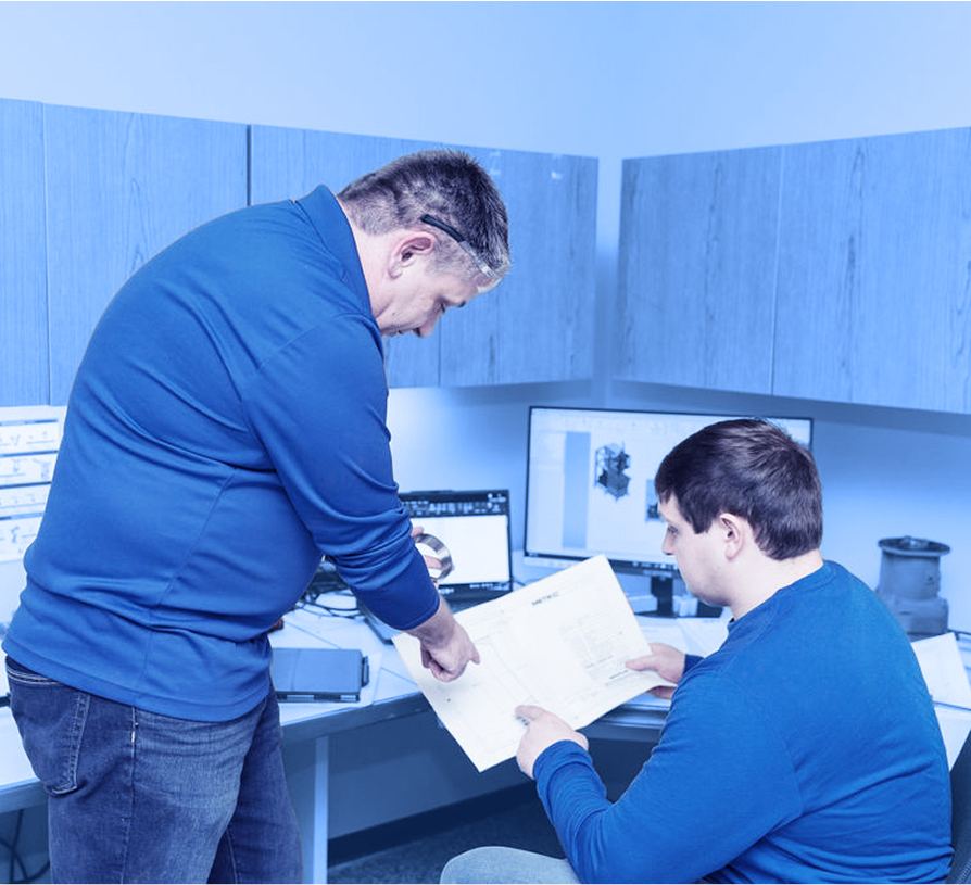 Two employees review printed engineering drawings at a desk with computer monitors displaying 3D component models.  