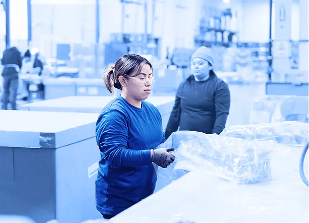 Worker wearing gloves and safety glasses placing plastic-wrapped parts onto a pallet inside a warehouse with boxes and other workers in the background.