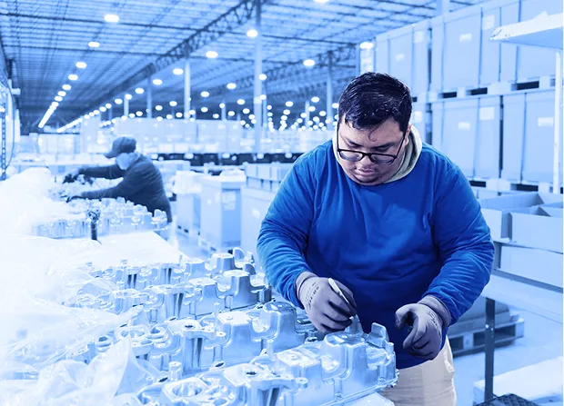Worker wearing gloves inspecting aluminum components on a worktable inside a warehouse with stacked boxes in the background.