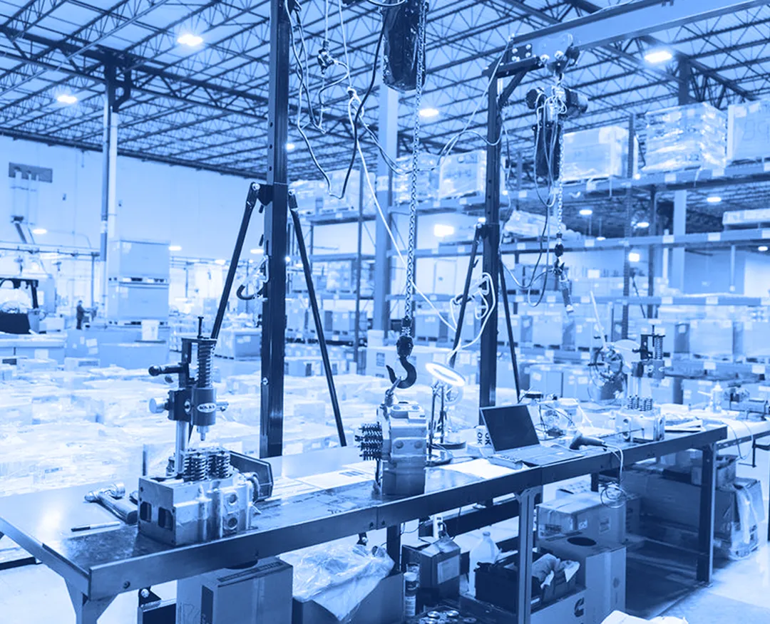 Workbench with suspended chain hoists, tools, and mechanical components set up inside a warehouse with storage racks and pallets in the background.
