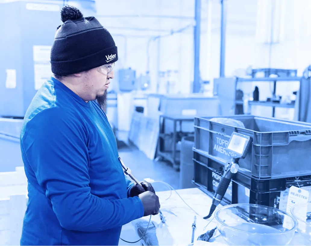 Worker wearing gloves, safety glasses, and a knit cap using a handheld tool at a workbench with plastic bins, containers, and equipment in a factory workspace.