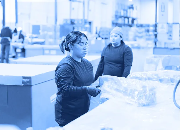 Worker wearing gloves and safety glasses placing plastic-wrapped parts onto a pallet inside a warehouse with boxes and other workers in the background.  
