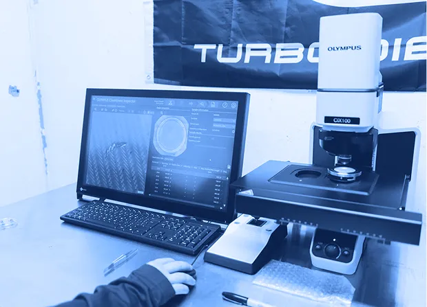 Computer workstation displaying inspection software beside a digital microscope on a lab bench with a hand resting near the keyboard.