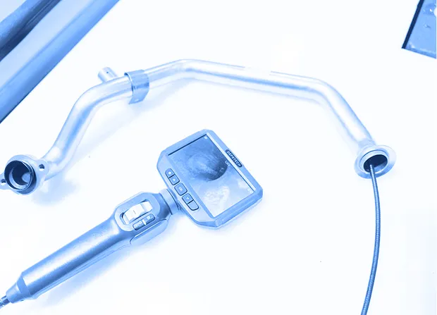 FA metal tube assembly on a work surface is inspected using a digital borescope, with the probe inserted into the tube and the internal surface visible on the handheld screen.