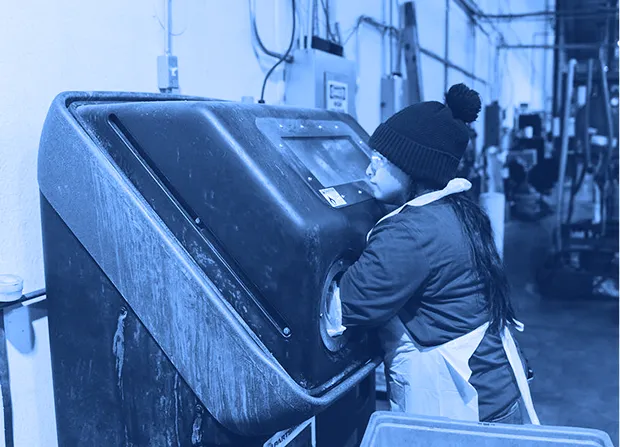 Worker wearing gloves, safety glasses, and a knit cap placing hands inside a blast cabinet while standing along a factory wall with equipment and bins nearby.