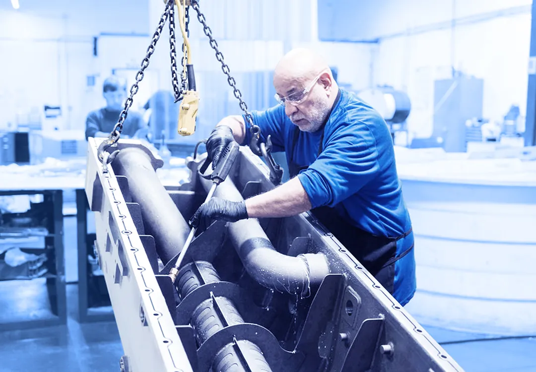 Worker wearing gloves and safety glasses using a handheld tool to work on large metal components suspended by chains inside a factory workspace.