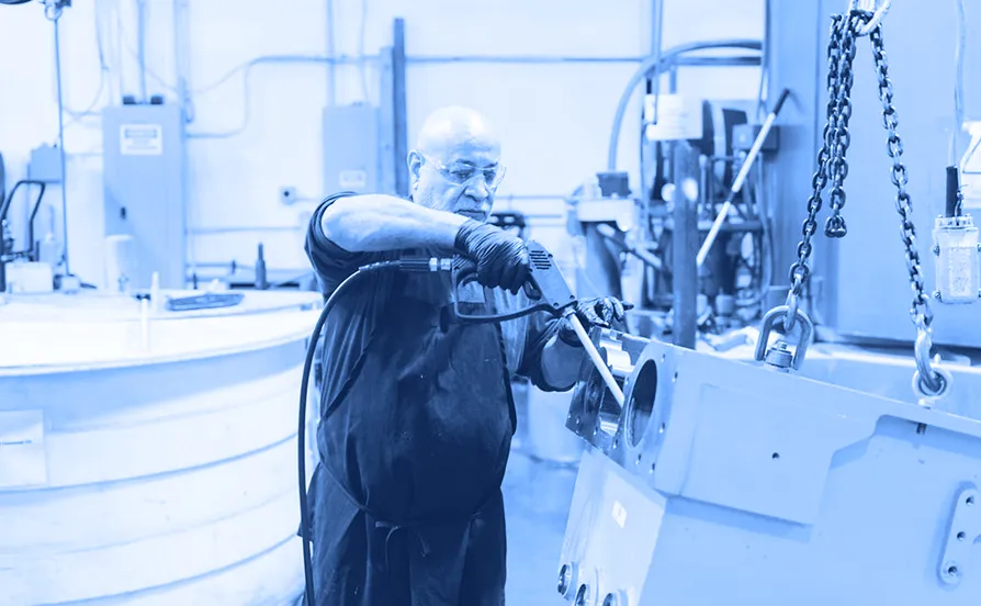 Worker wearing gloves and safety glasses using a handheld washing tool on a large metal component suspended by chains inside a factory workspace.