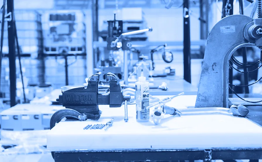 Workbench with a metal vise, hand tools, fasteners, and a bottle of adhesive arranged on a tabletop inside a factory area.